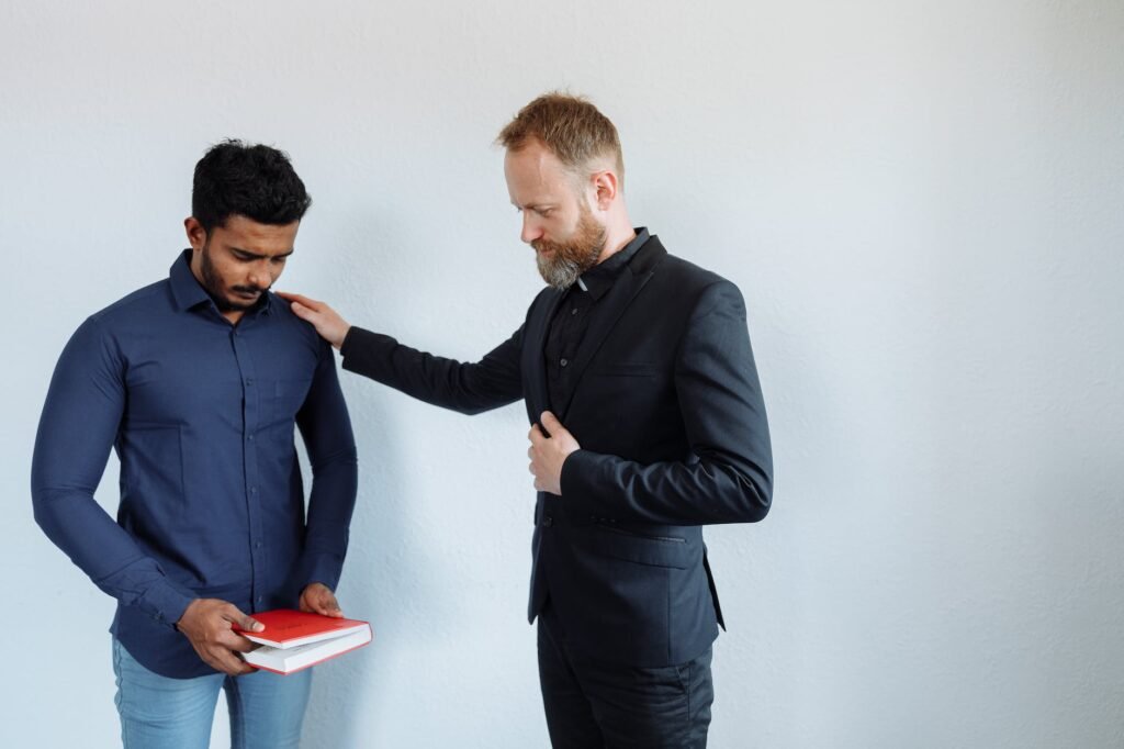 man with closed eyes holding a red book while man in black suit holding hand on his shoulder