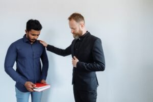man with closed eyes holding a red book while man in black suit holding hand on his shoulder